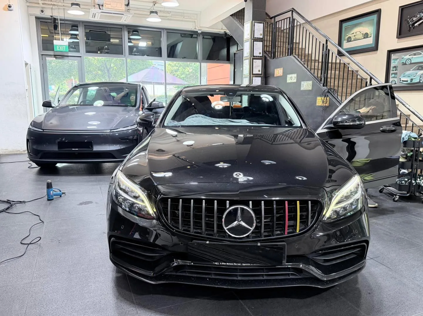 A black Mercedes-Benz with a distinctive grille in the foreground, with a dark Tesla and another car behind it.