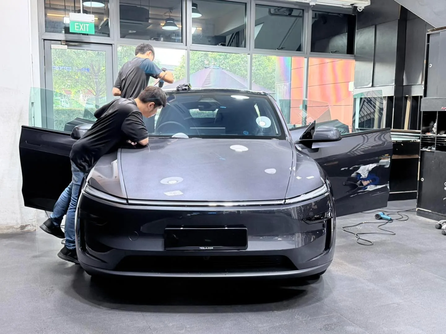 A dark-coloured car with two technicians applying film to the side windows.