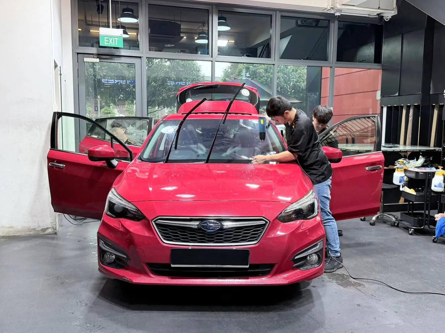 A bright red Subaru Impreza being treated, doors open, with technician working on windows.