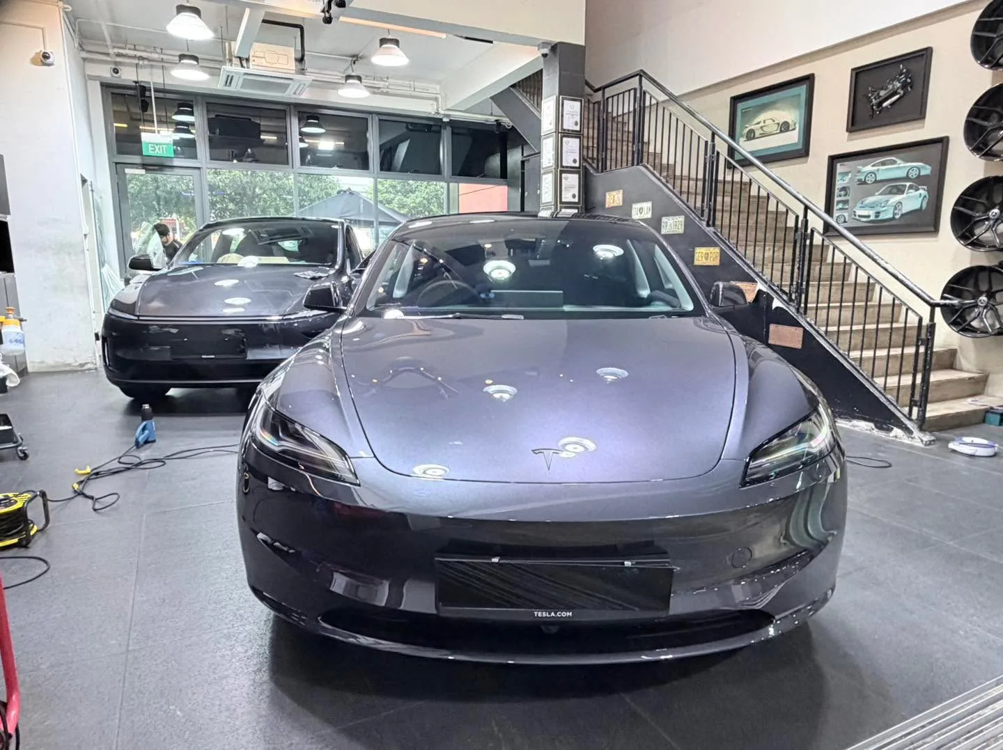 A dark grey Tesla sedan on the workshop floor with another dark vehicle behind.
