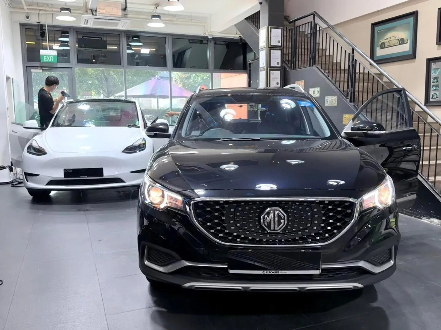 A black MG sedan with distinctive grille parked in the workshop next to a white Tesla.
