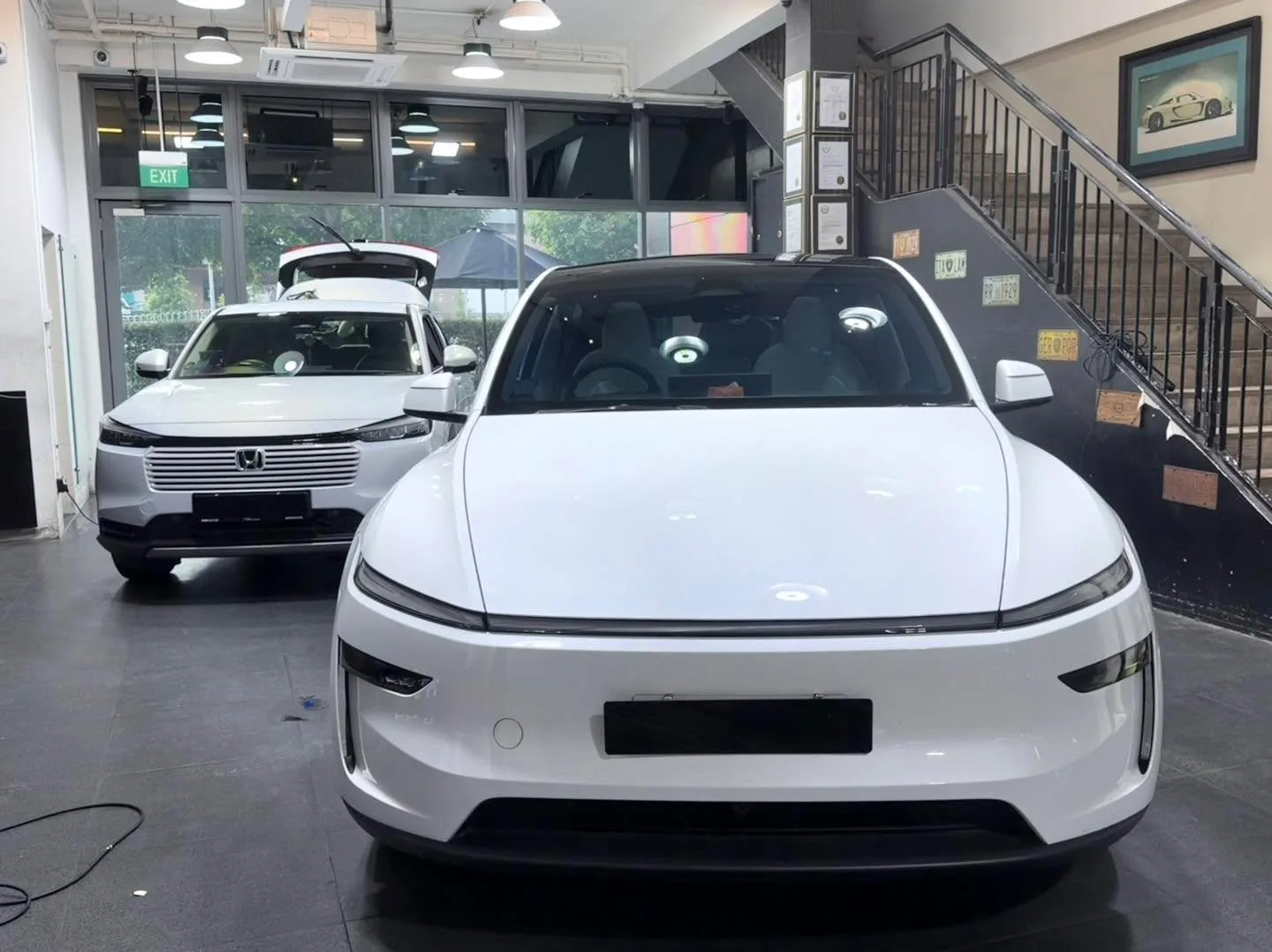 A white Tesla sedan on the workshop floor with a white Honda SUV behind it.