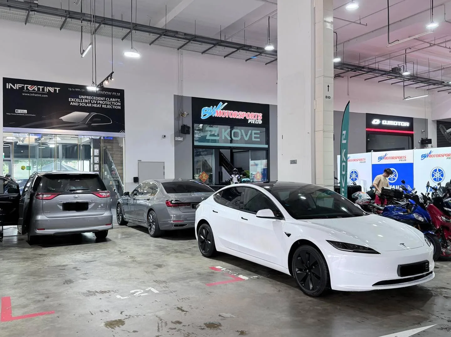 A white Tesla and blue sedan in a workshop bay under fluorescent lighting.