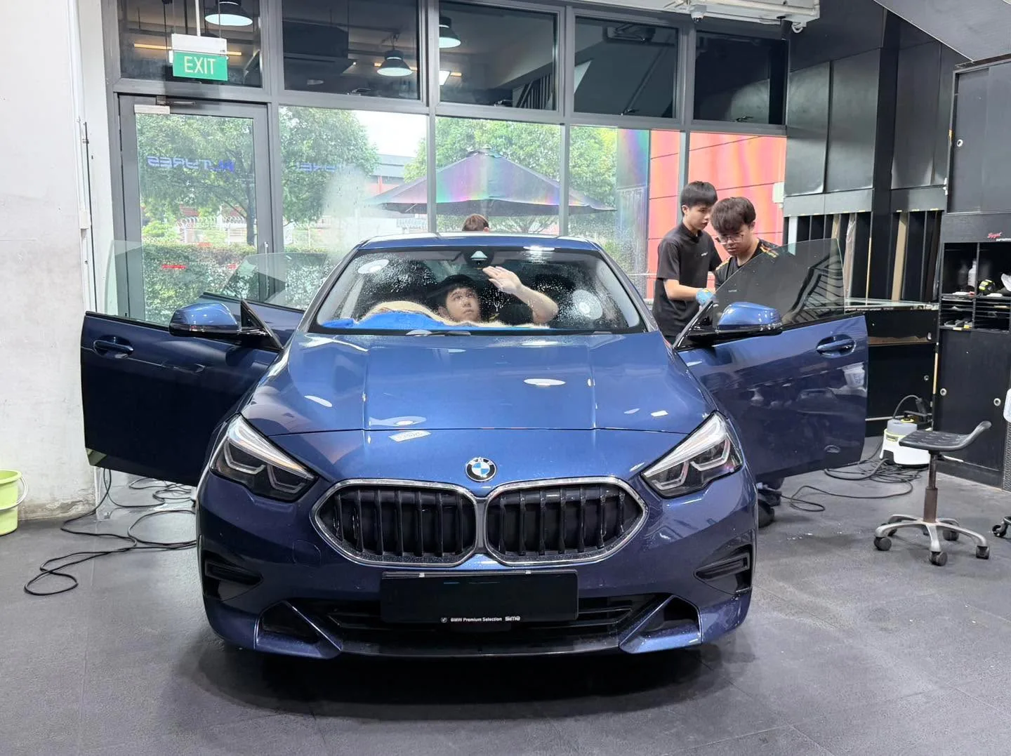 A blue saloon car with workers visible inside applying window tint to the interior.