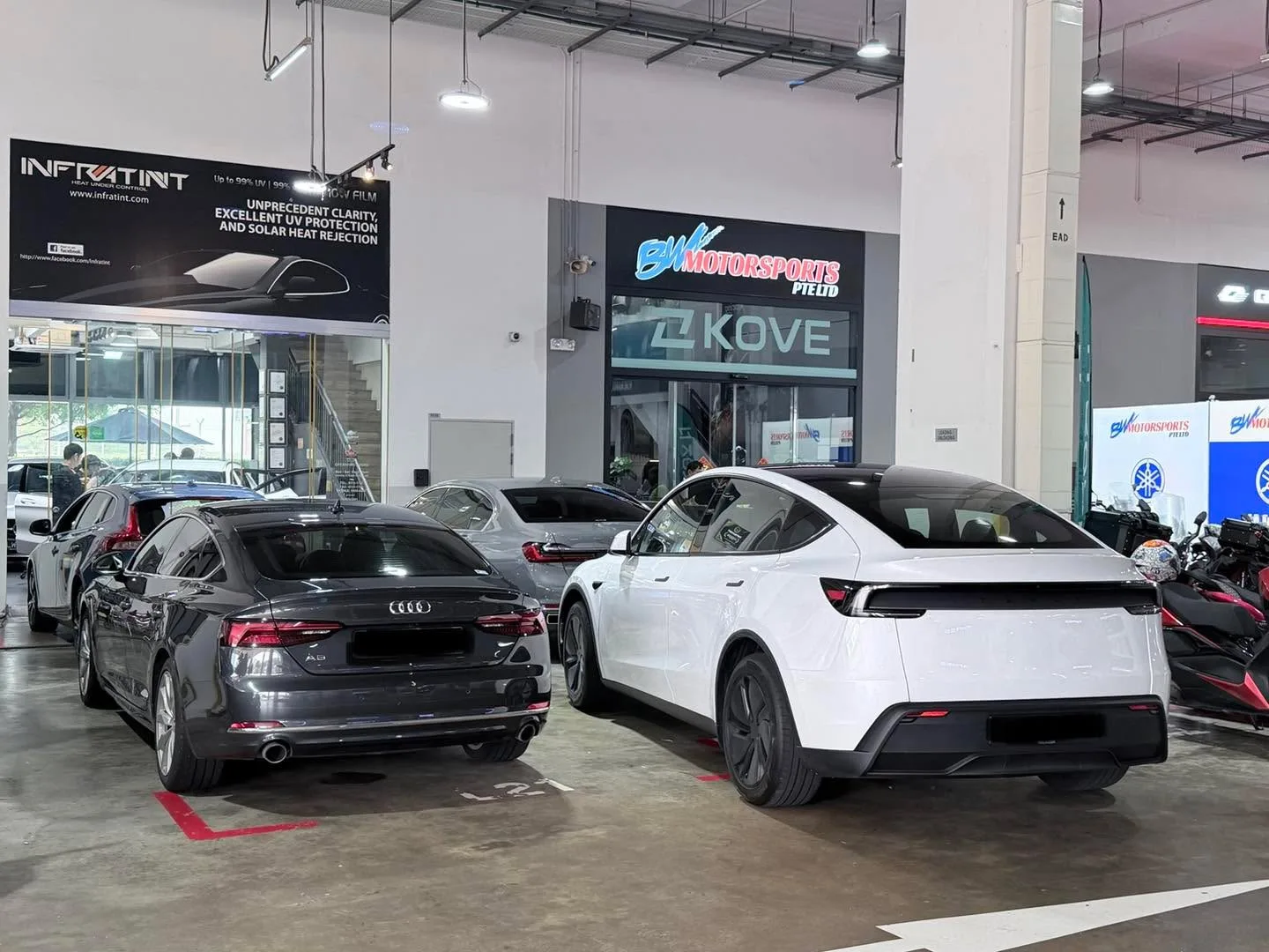 A dark grey Audi A5 sedan and a white Tesla with hatchback body parked side by side in a workshop bay.