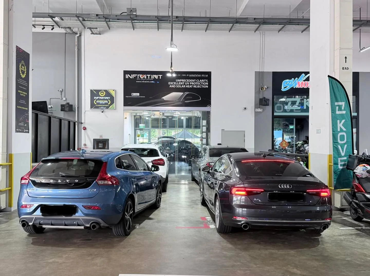 A blue hatchback and a dark Audi sedan viewed from behind in a workshop, with the storefront visible.