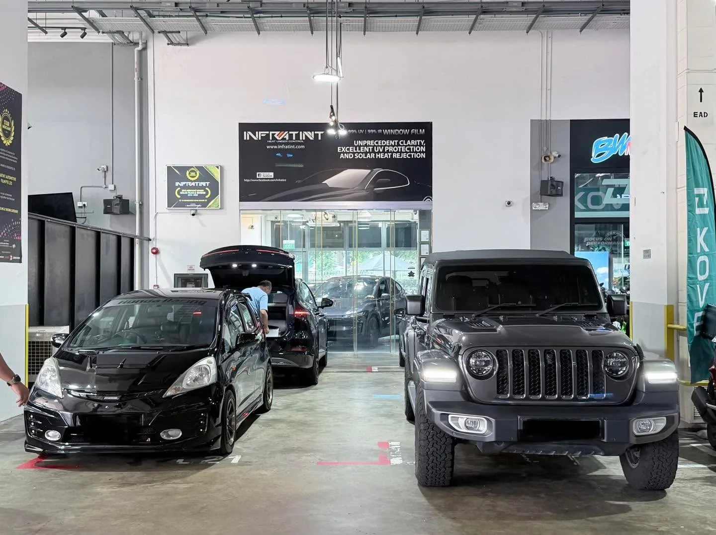 A black hatchback with open door doors and a dark Jeep SUV with round headlights inside the workshop.