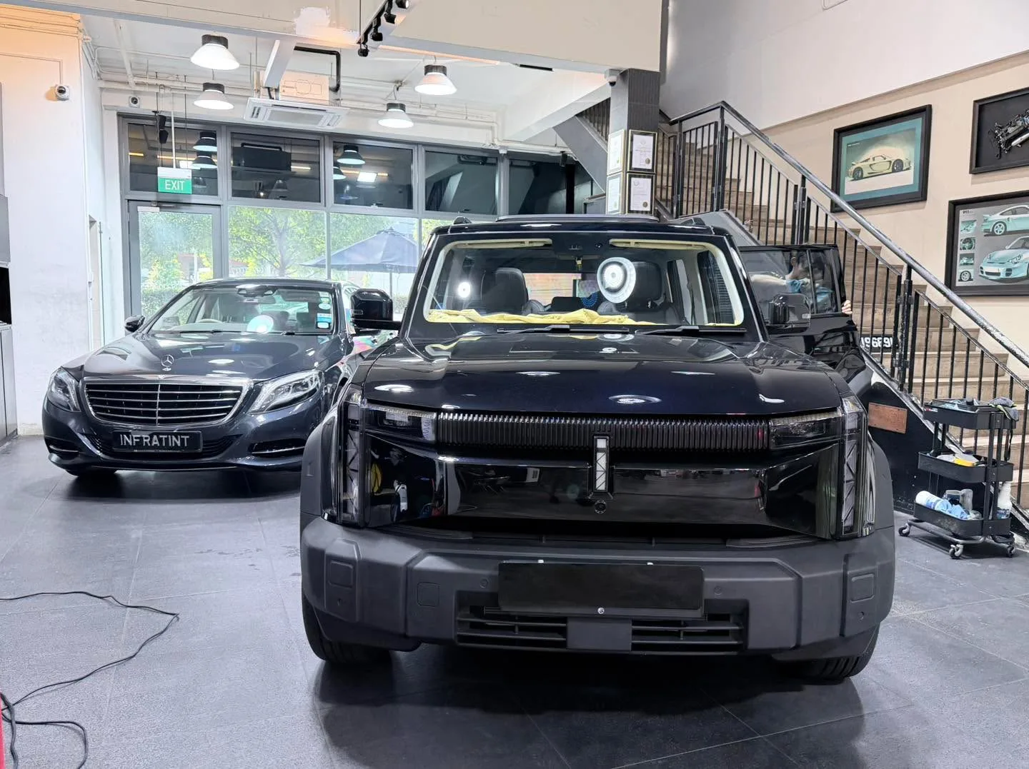 A black SUV with its bonnet open, front doors ajar, in an indoor workshop bay. A dark saloon is visible behind it.