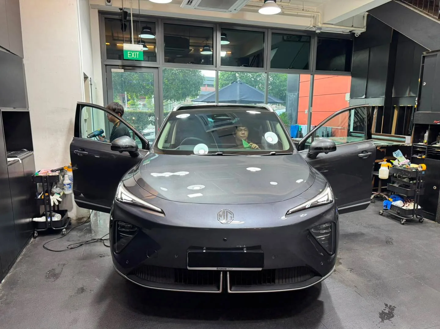 A grey compact electric SUV with doors open, parked in an indoor workshop bay with work equipment nearby.