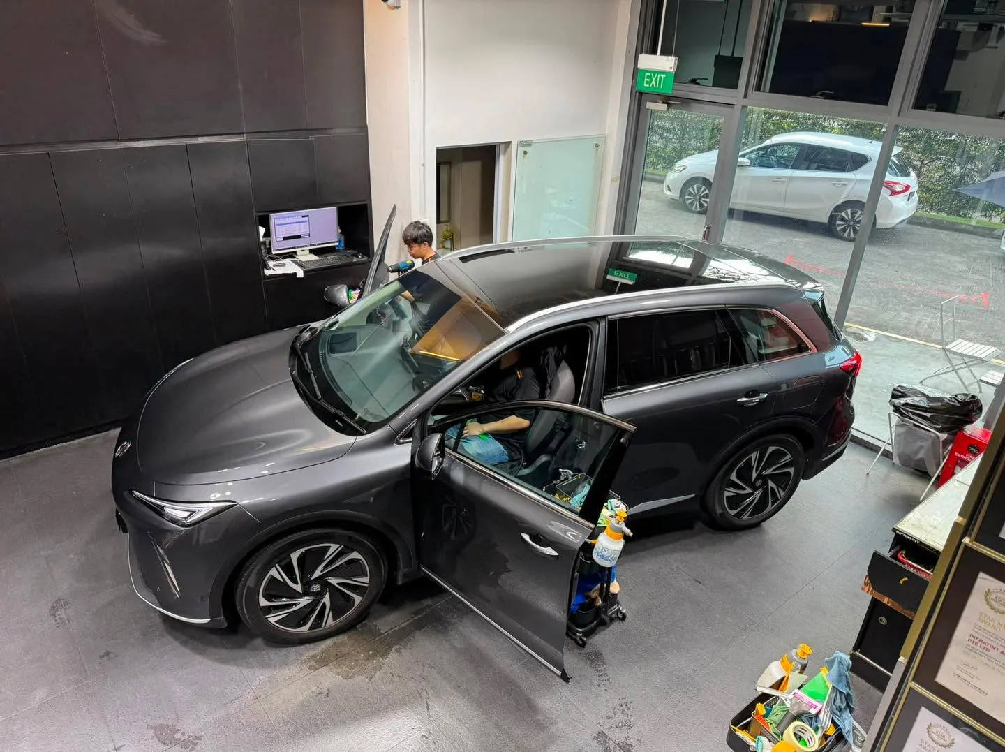 A dark grey SUV with doors and bonnet open in an indoor workshop. A white car is visible outside through large windows.