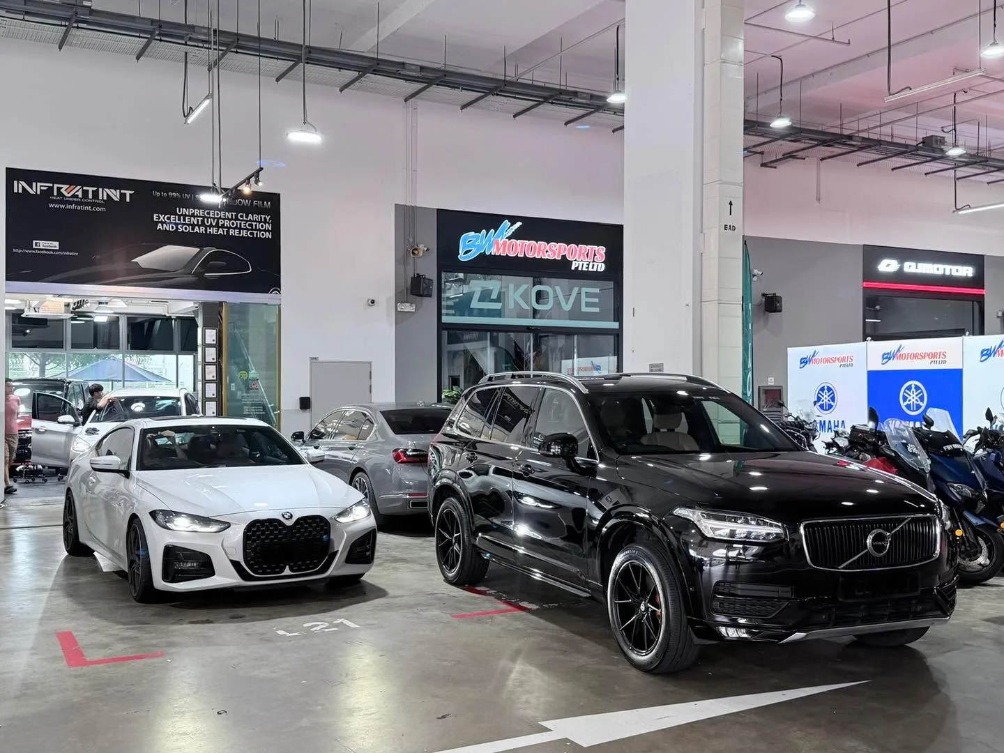 A white BMW and a black Volvo SUV parked in a workshop beside display signage.