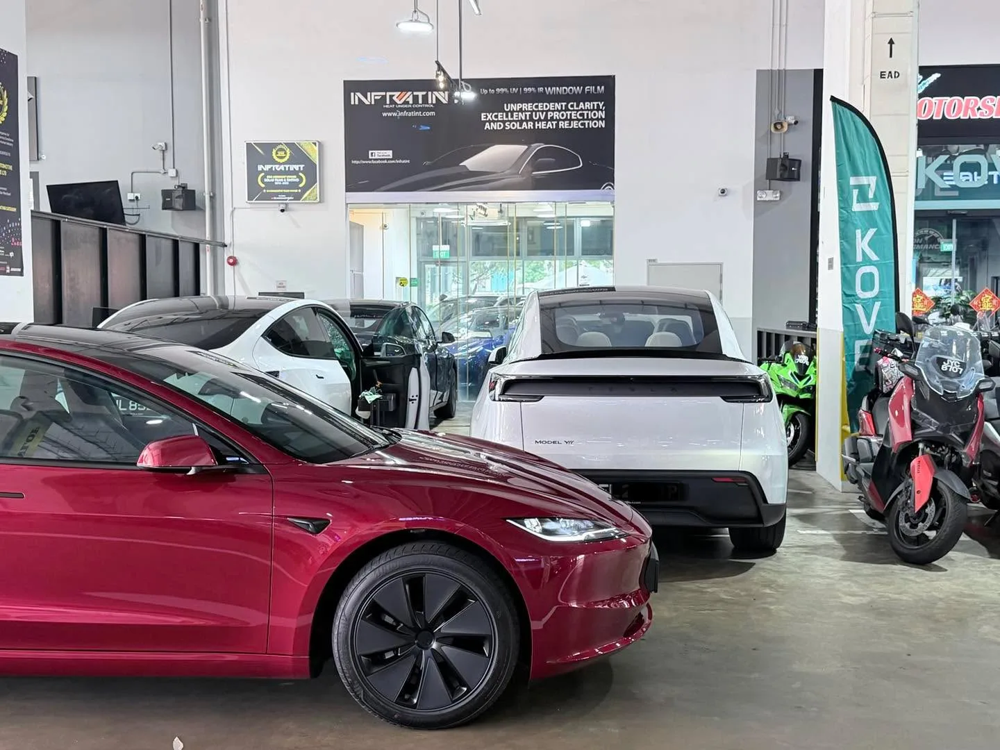 A red Tesla Model Y L and a white Lucid vehicle parked indoors next to the workshop's signage.