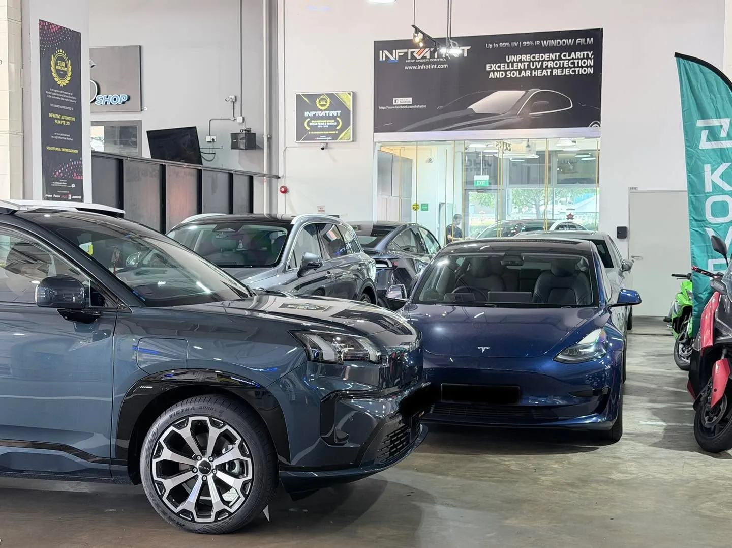 A grey Tesla Model Y, a blue Tesla Model 3 Highland, and other vehicles in the workshop bay.