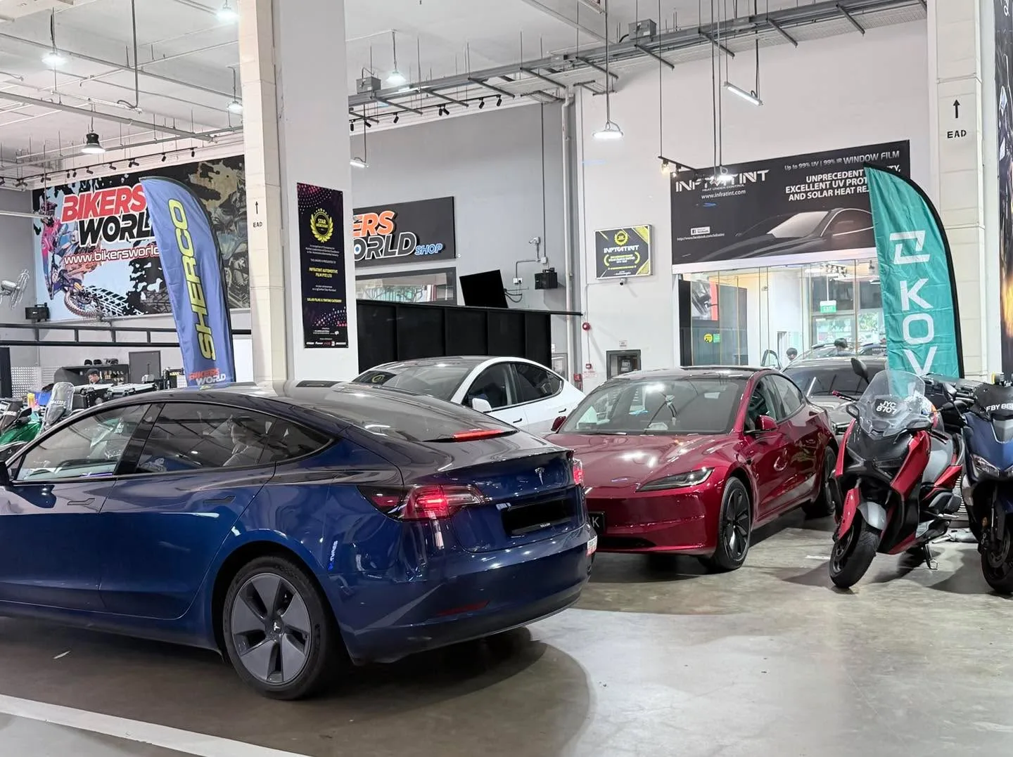 A blue Tesla Model 3 in the foreground and a maroon Tesla Model Y in the background, parked indoors.