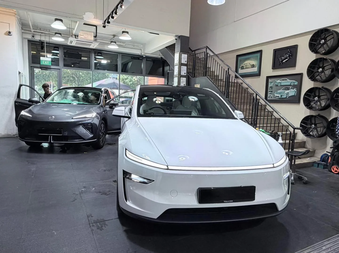A white Tesla Model Y and a dark blue Tesla Model 3 parked indoors with a person visible.