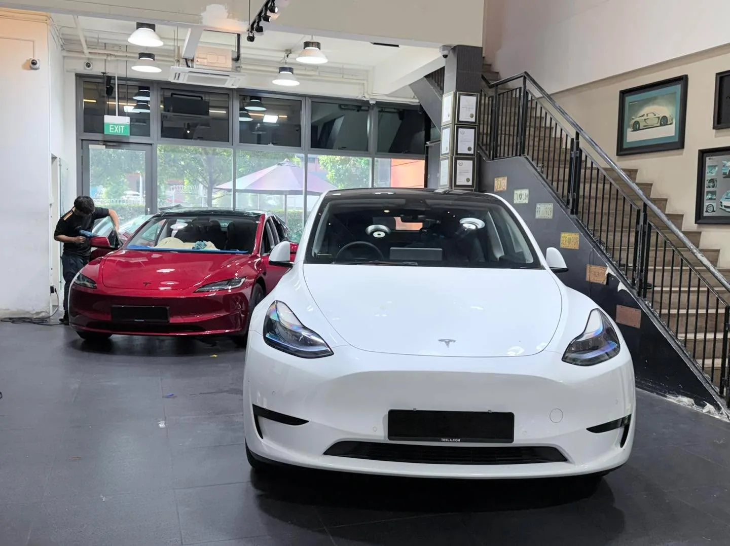 A white Tesla Model Y in the foreground and a red Tesla Model Y beside a Mazda CX-5 with an open boot.