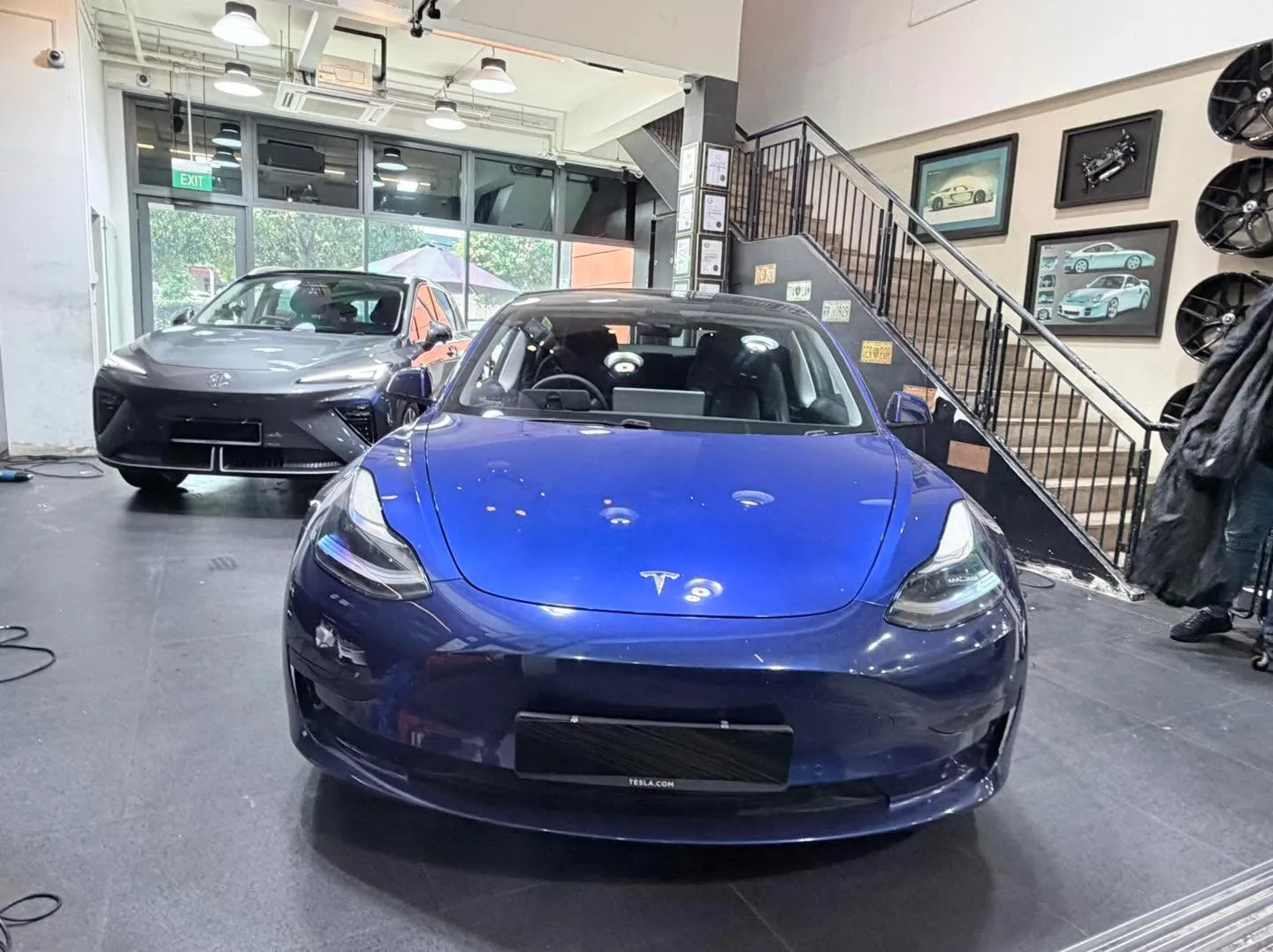 A blue Tesla Model 3 in the foreground with other vehicles visible behind it in the workshop.
