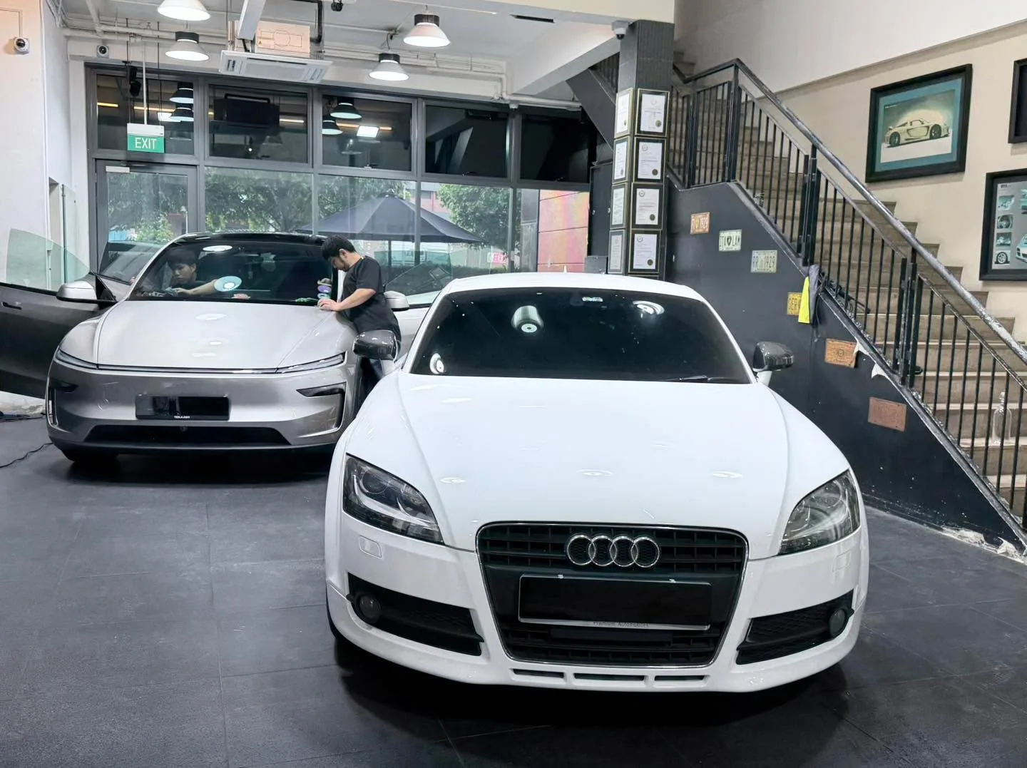 White Audi TT and silver Tesla SUV facing each other in workshop interior.