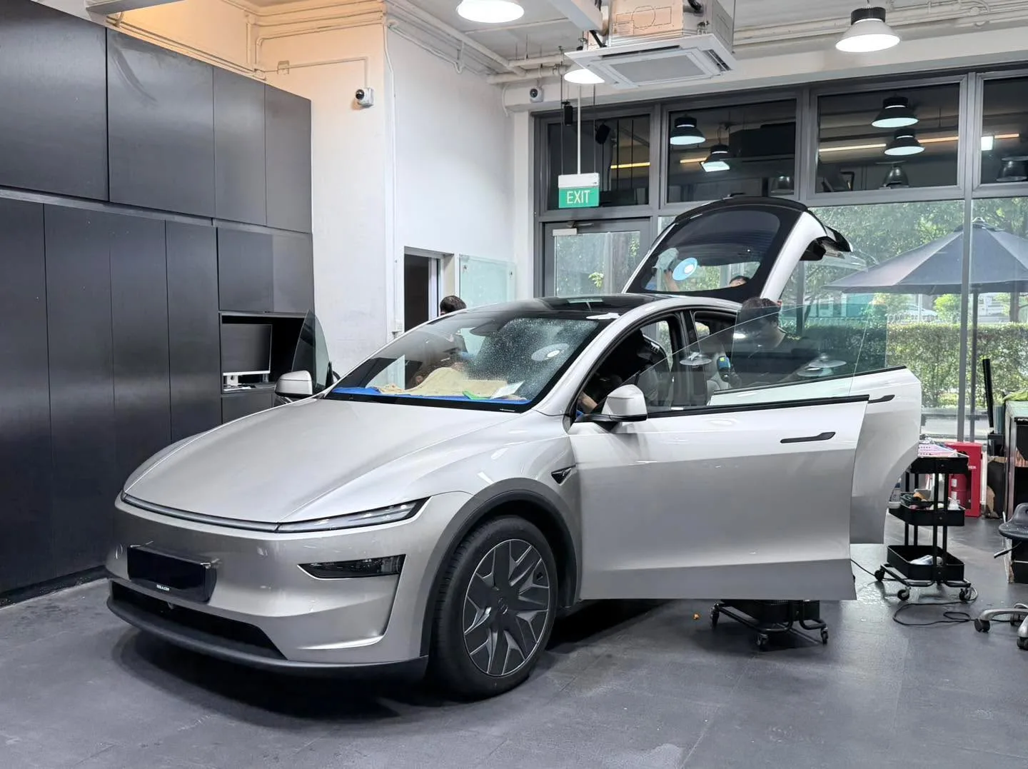 Silver Tesla SUV with driver door open, technician working on window in workshop.