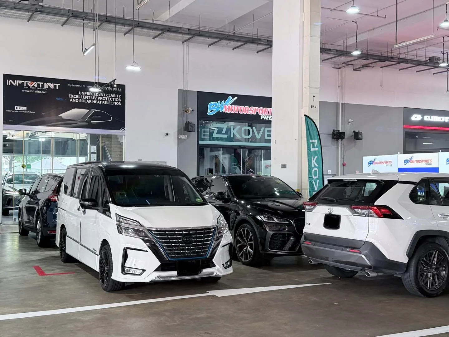 A white Nissan Serena and black Kia Niro parked in a workshop bay with other vehicles.