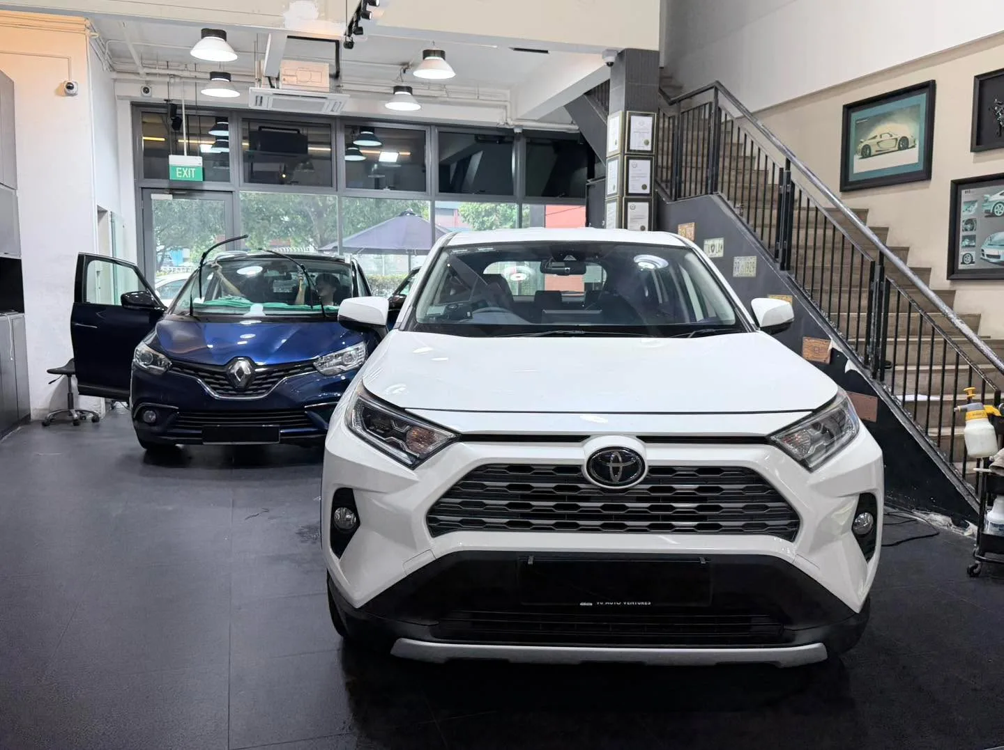A white Nissan Serena and blue Renault Grand Scenic in a workshop with a staircase.