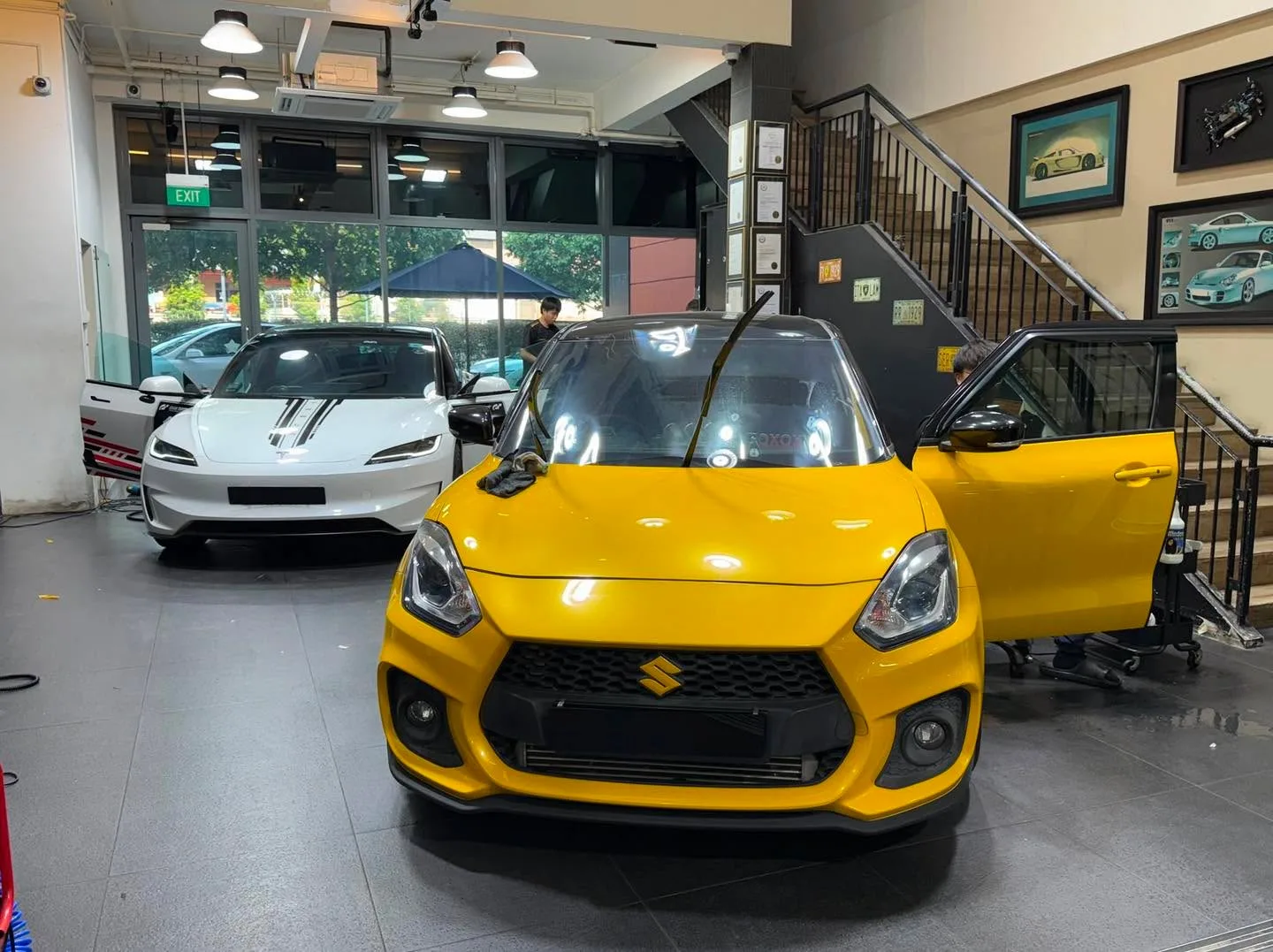 Yellow Suzuki Swift with open door and white Tesla in workshop.