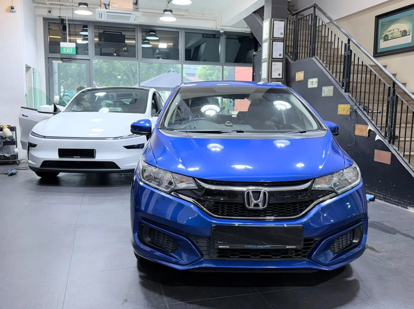 A bright blue Honda Jazz hatchback viewed from the front, with a white Tesla behind.