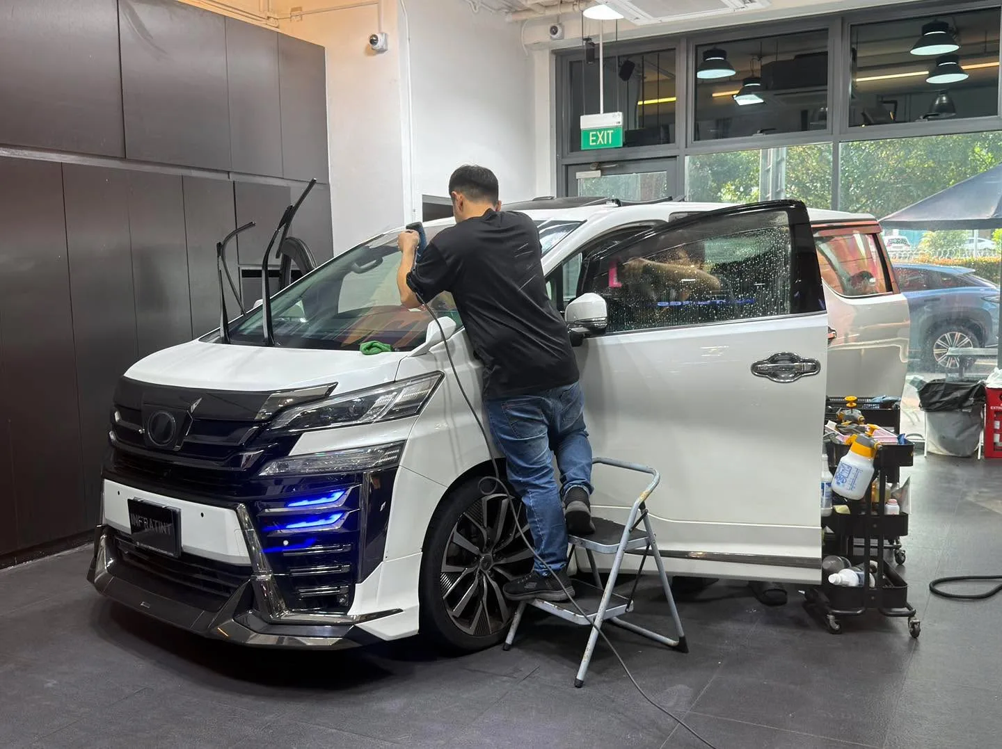 A grey saloon with gull-wing doors open, undergoing window film application.
