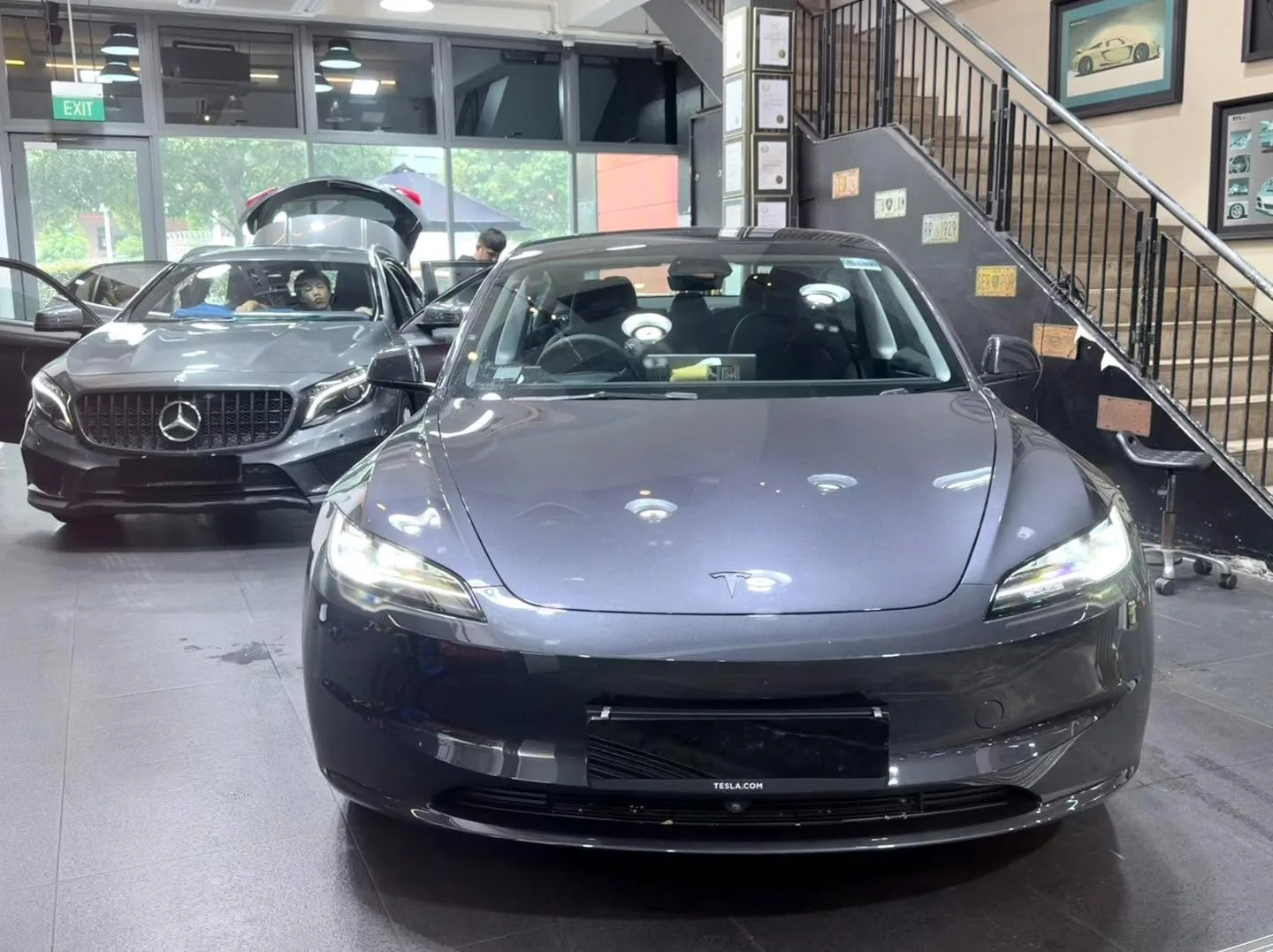 A black Tesla sedan in the foreground with doors open, and a dark-coloured Mercedes-Benz behind.