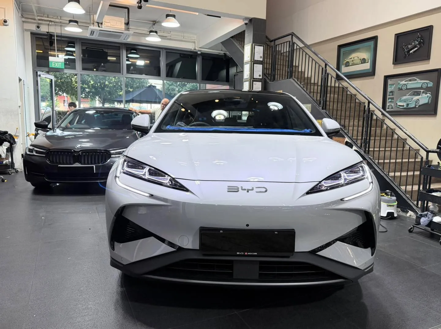 A white Tesla Model Y sedan with a black BMW sedan visible behind it in an indoor workshop.