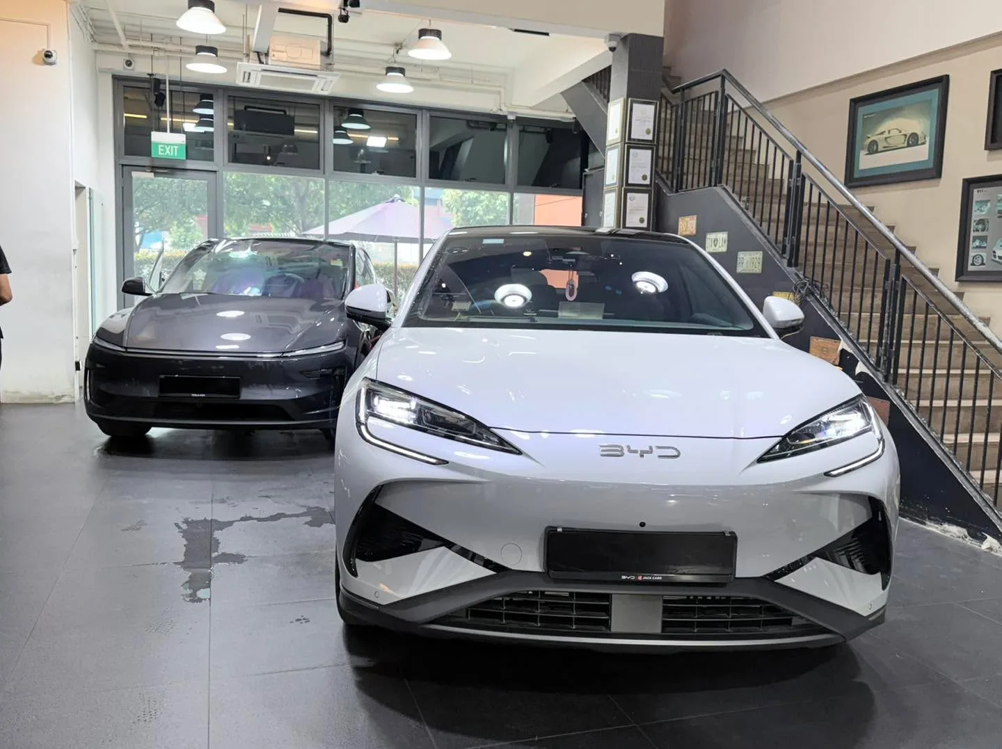 A black Tesla sedan and a white BYD sedan parked indoors with glass storefront visible.