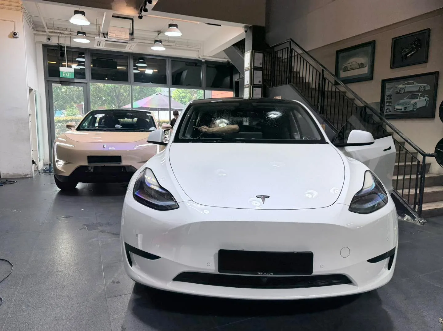A white Tesla Model Y parked indoors with a beige car visible behind it.