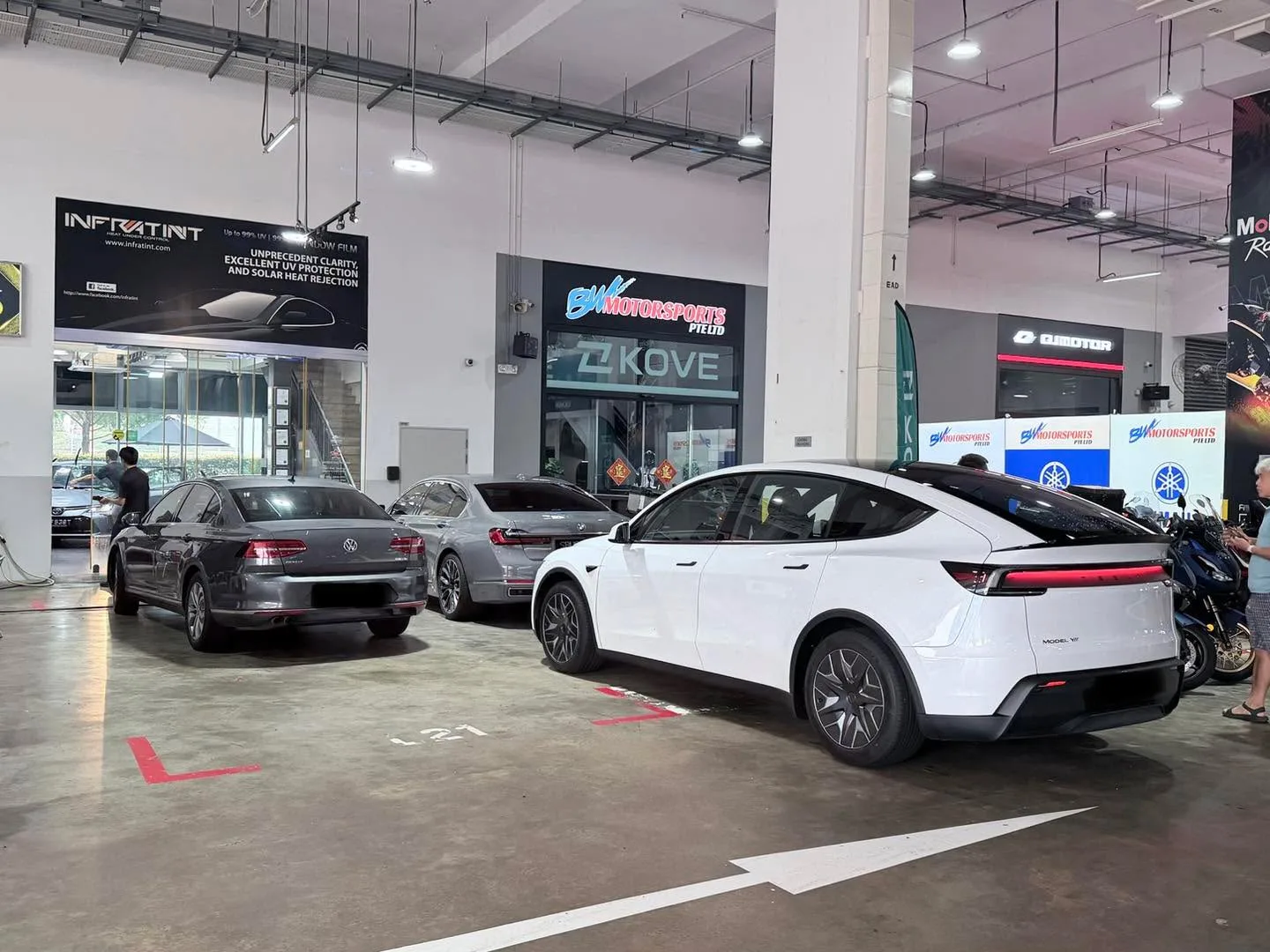 A white sedan and a dark grey sedan in a workshop bay with industrial ceiling and signage visible.