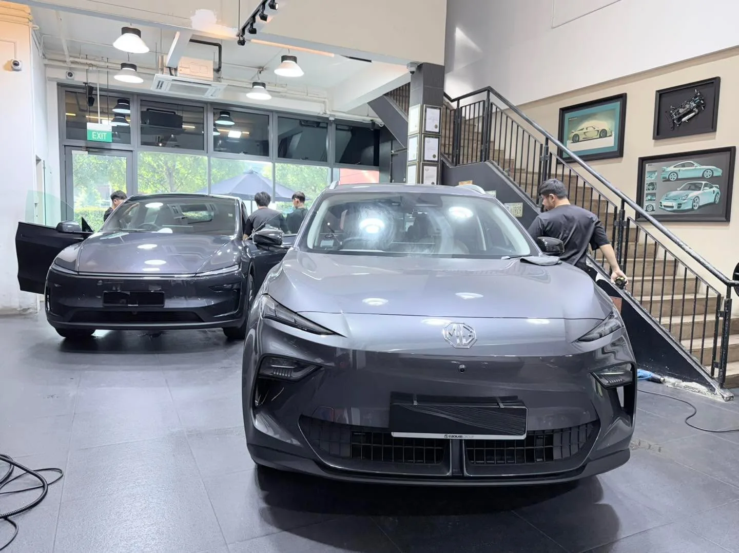 A dark grey electric sedan and a white electric sedan being tinted by workshop staff.