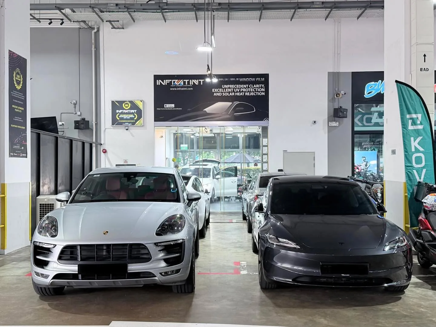 A white Porsche SUV and a dark Tesla sedan parked side by side in the workshop.