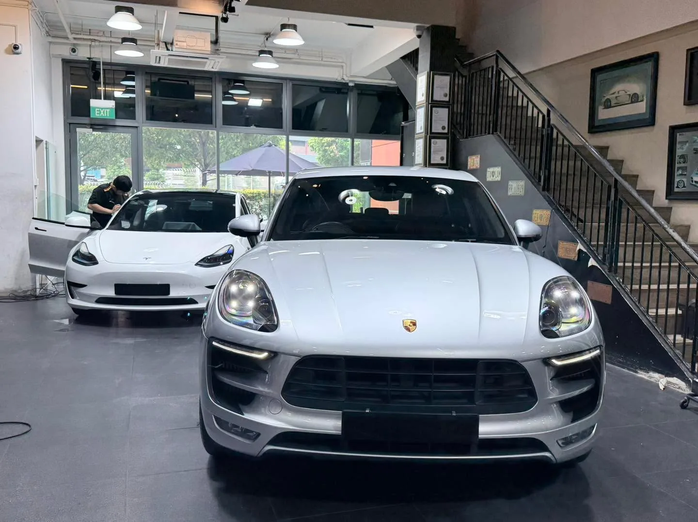 A silver Porsche SUV in the foreground with a white Tesla sedan behind it inside the workshop.