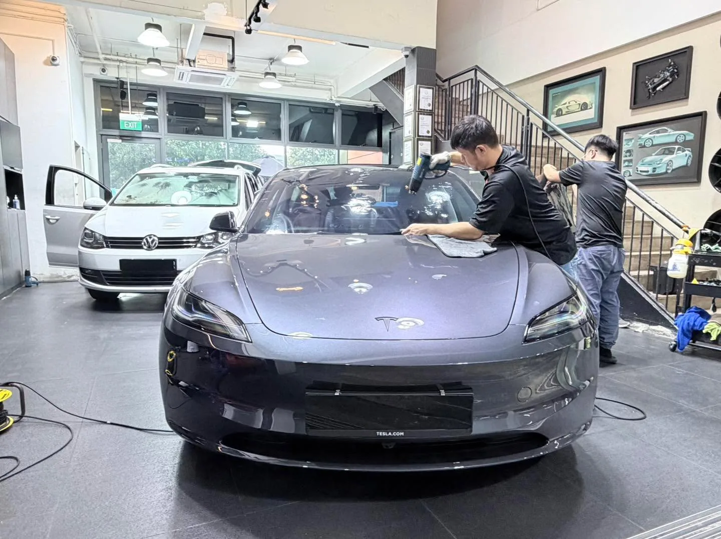 Two technicians working on a dark Tesla sedan with doors open, with a white Volkswagen hatchback visible on the left.