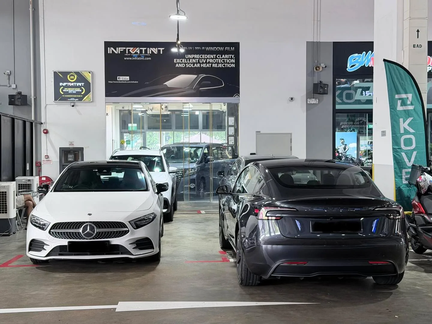 A white Mercedes-Benz and a dark Tesla sedan parked in the workshop showroom.