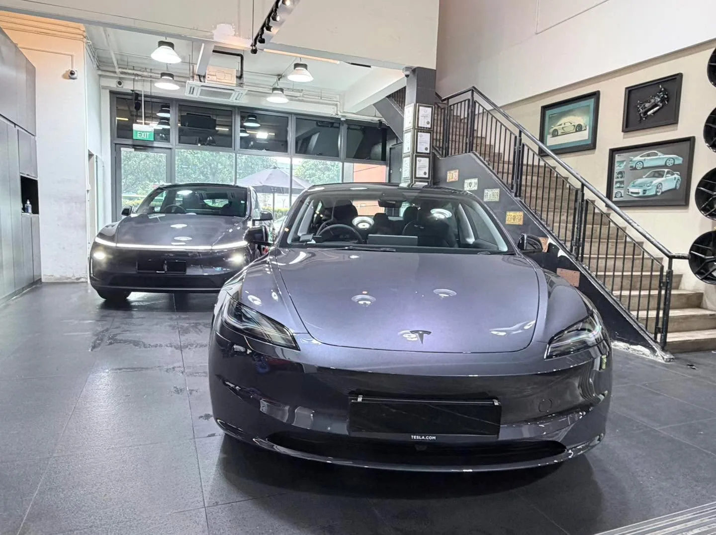 A black Tesla sedan in the foreground with a darker sedan behind it inside the workshop.