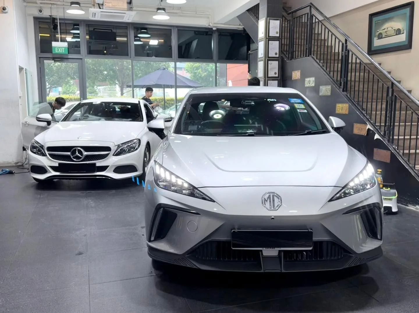 A white Mercedes-Benz sedan in the foreground and a white MG compact hatchback behind it inside the workshop.