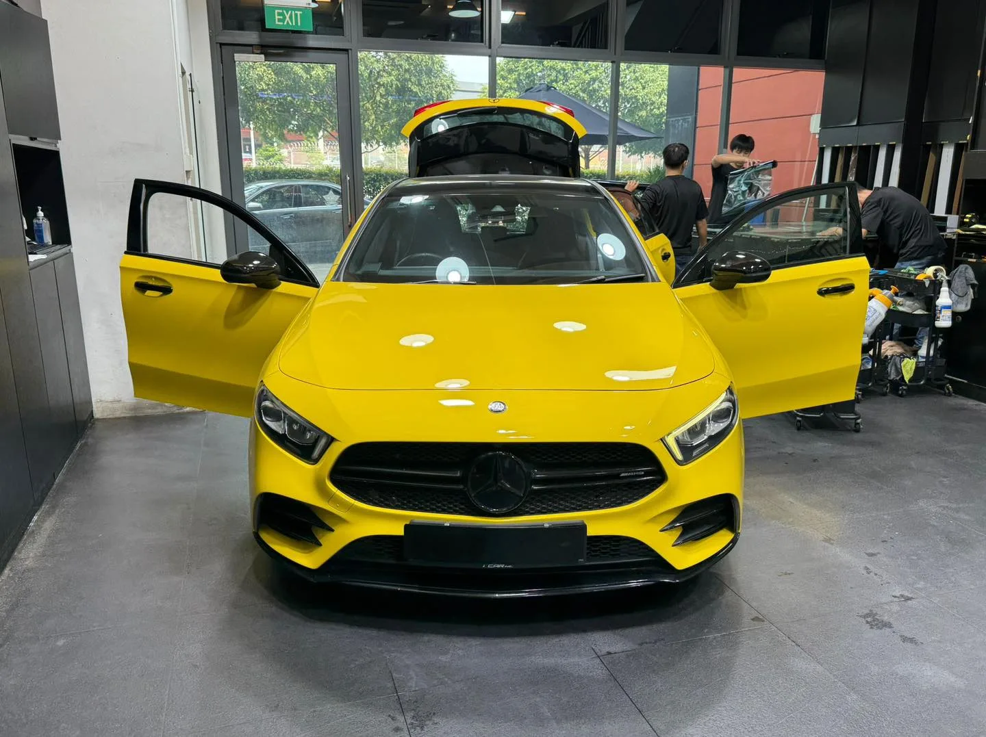 A bright yellow Mercedes-Benz compact car with doors open, parked inside the workshop with technicians working nearby.