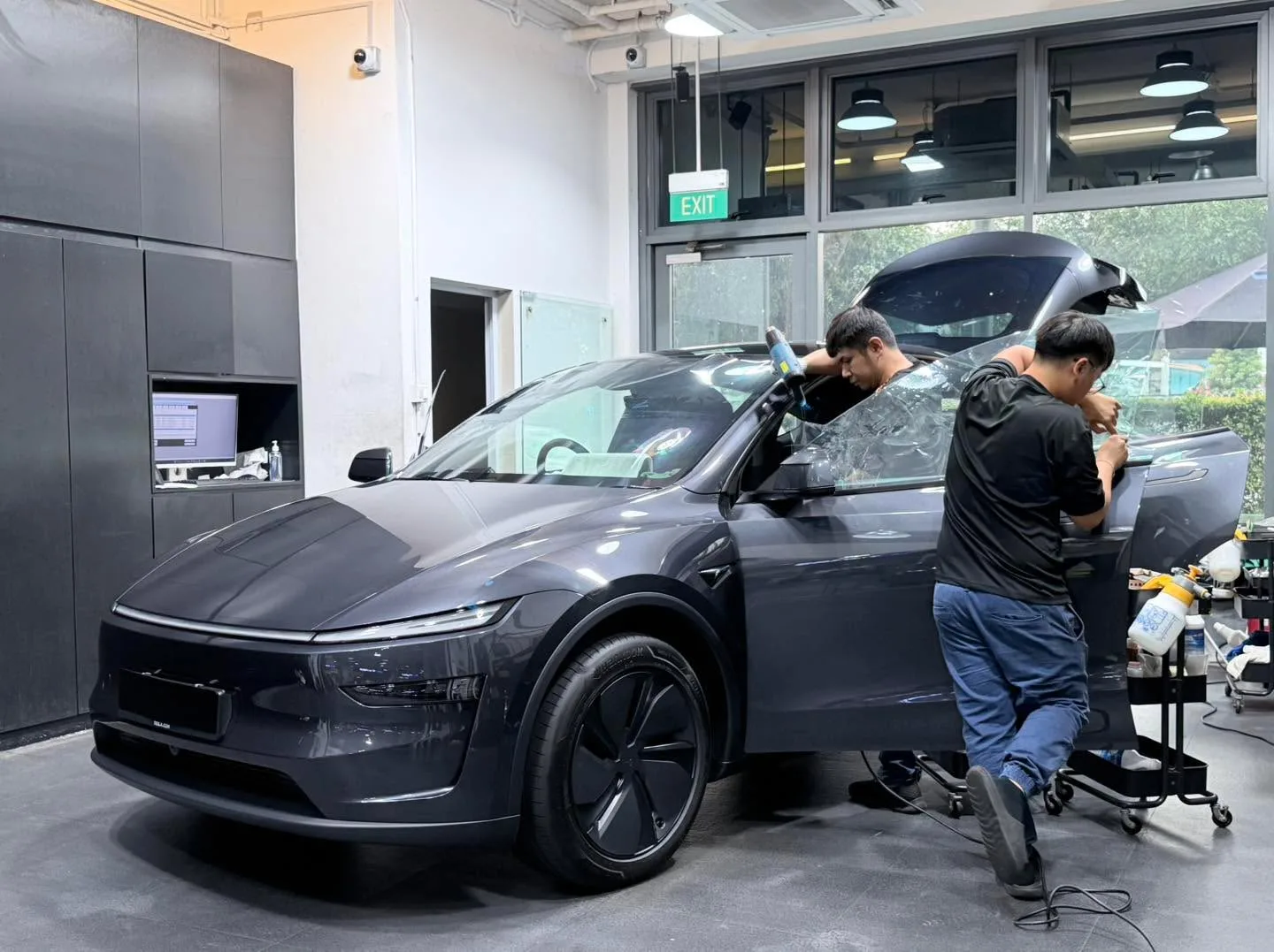A dark grey electric SUV with gullwing door open, technician applying window film.