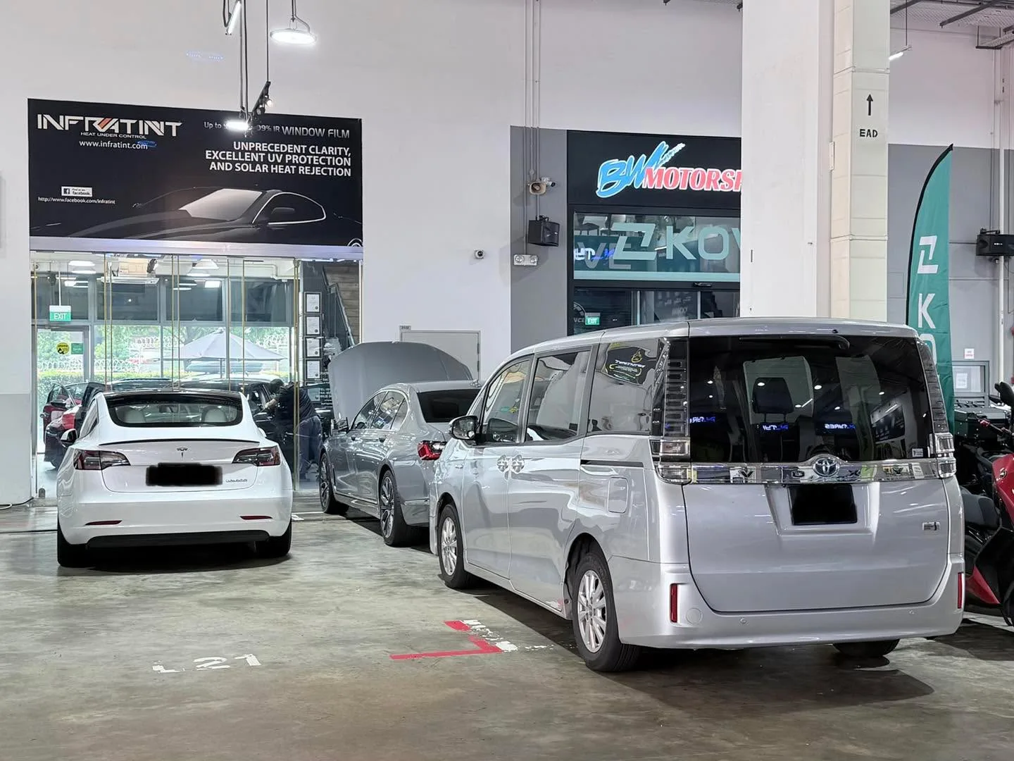 White Tesla Model 3 and silver Toyota Noah in workshop bay with Infratint signage visible.