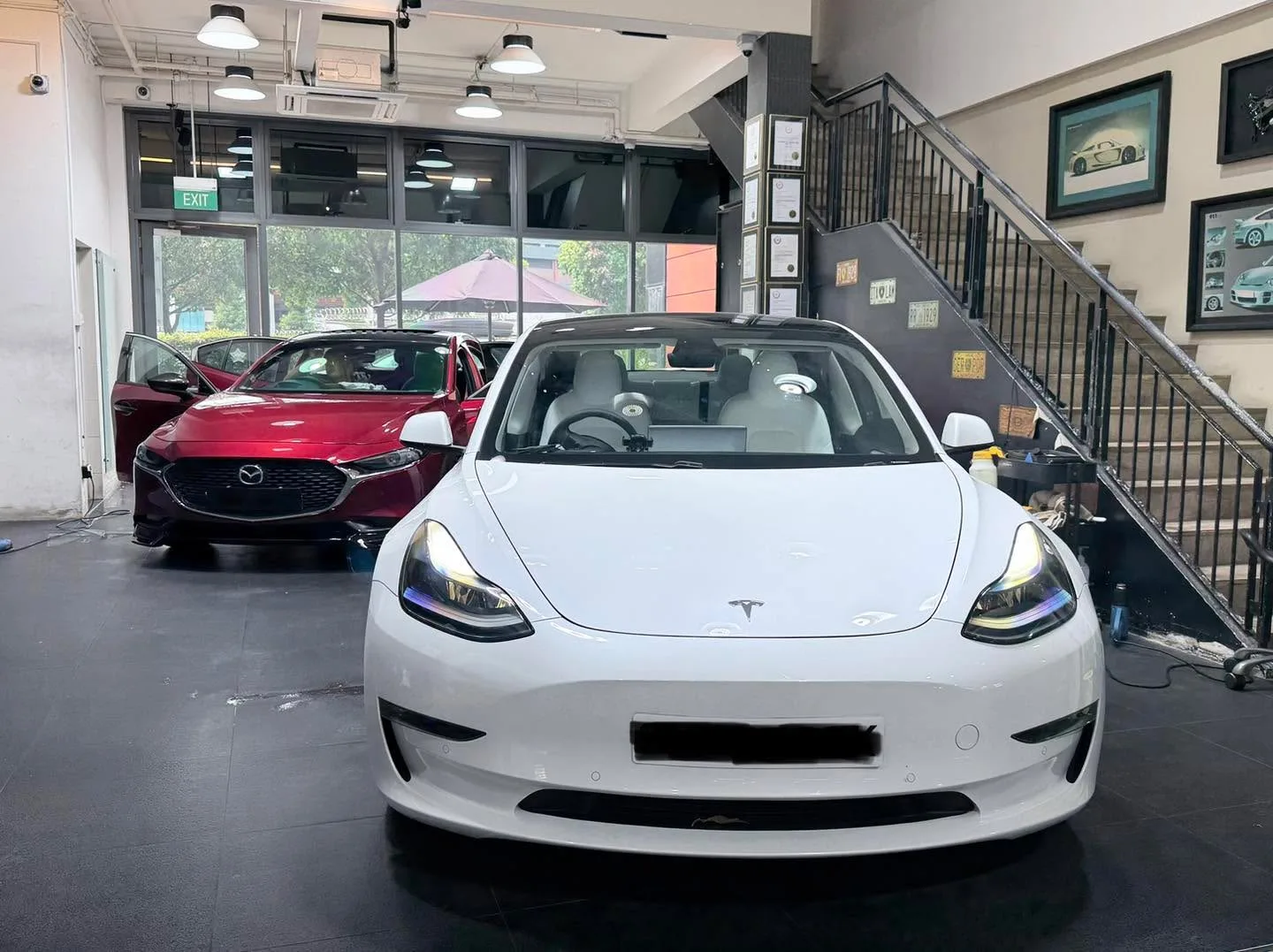 White Tesla Model 3 and red Mazda 3 in workshop with stairs behind.