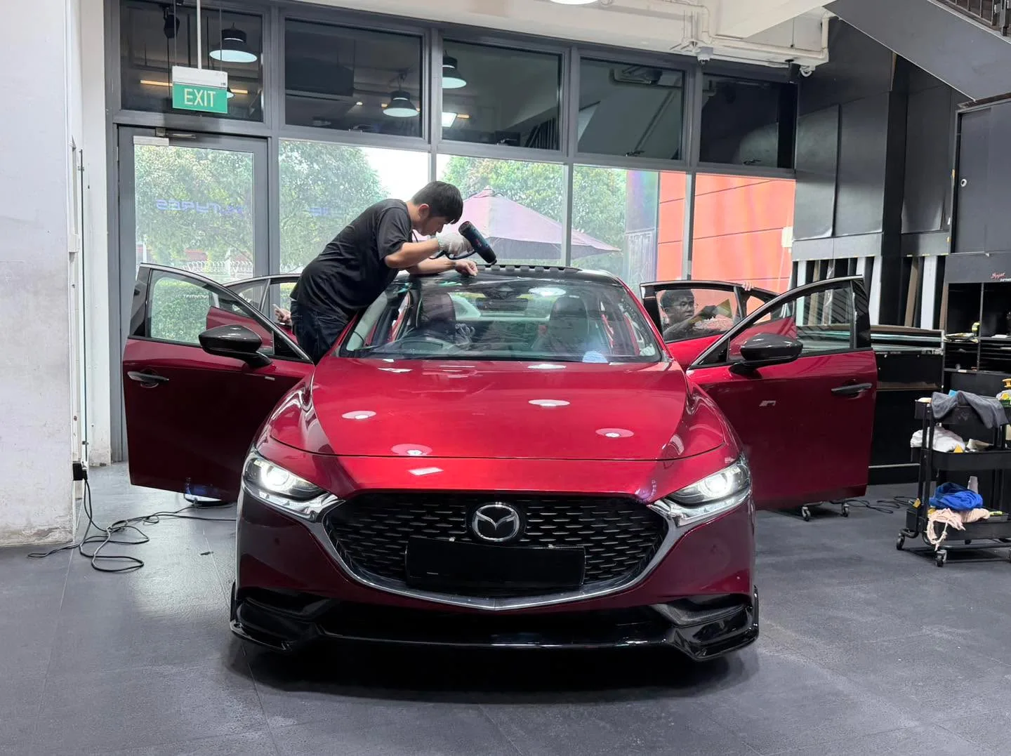 Red Mazda 3 sedan with open doors, technician working on window film application.