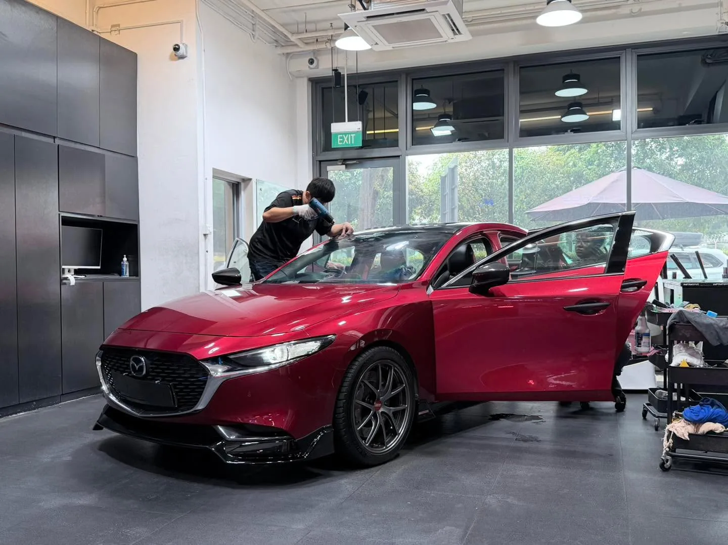Red Mazda 3 sedan with both front doors open, technician working on side window.
