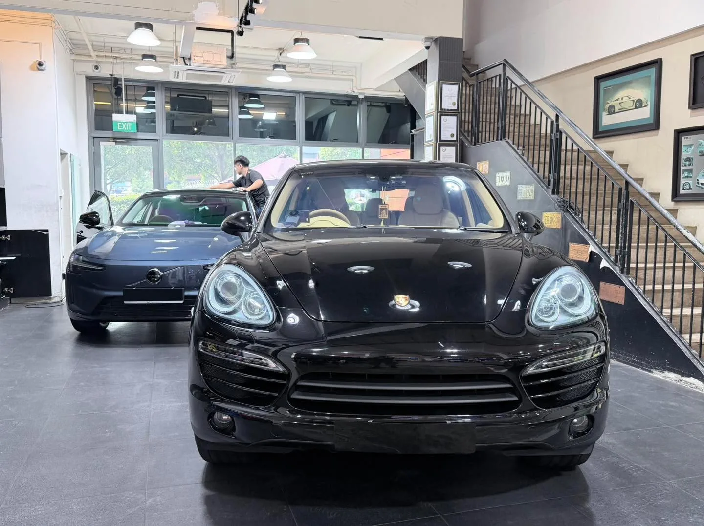 A black Porsche sports car parked indoors with a blue sedan visible in the background.