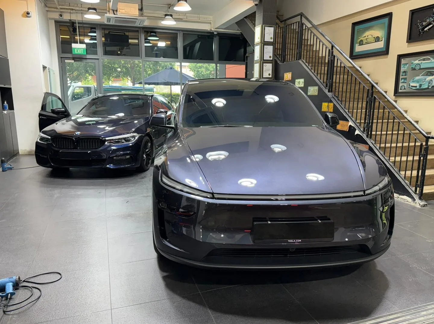 A dark grey Tesla electric sedan parked in a bright workshop with exposed lighting.