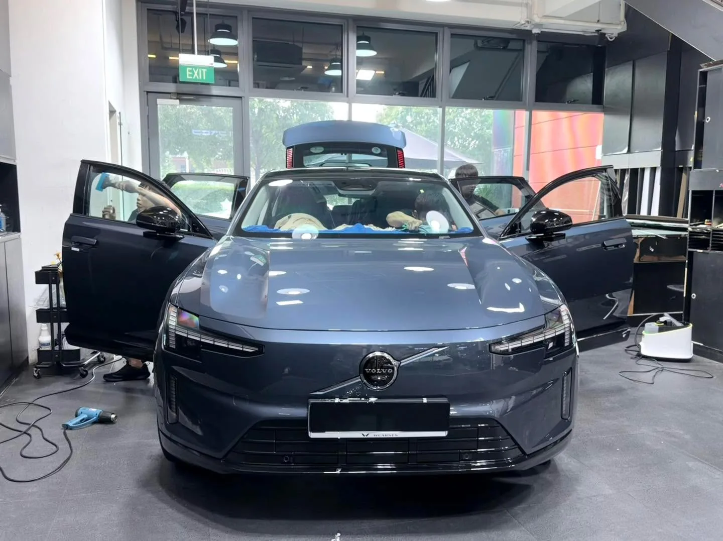 A dark grey Volvo electric sedan with both doors open, parked indoors under workshop lights.