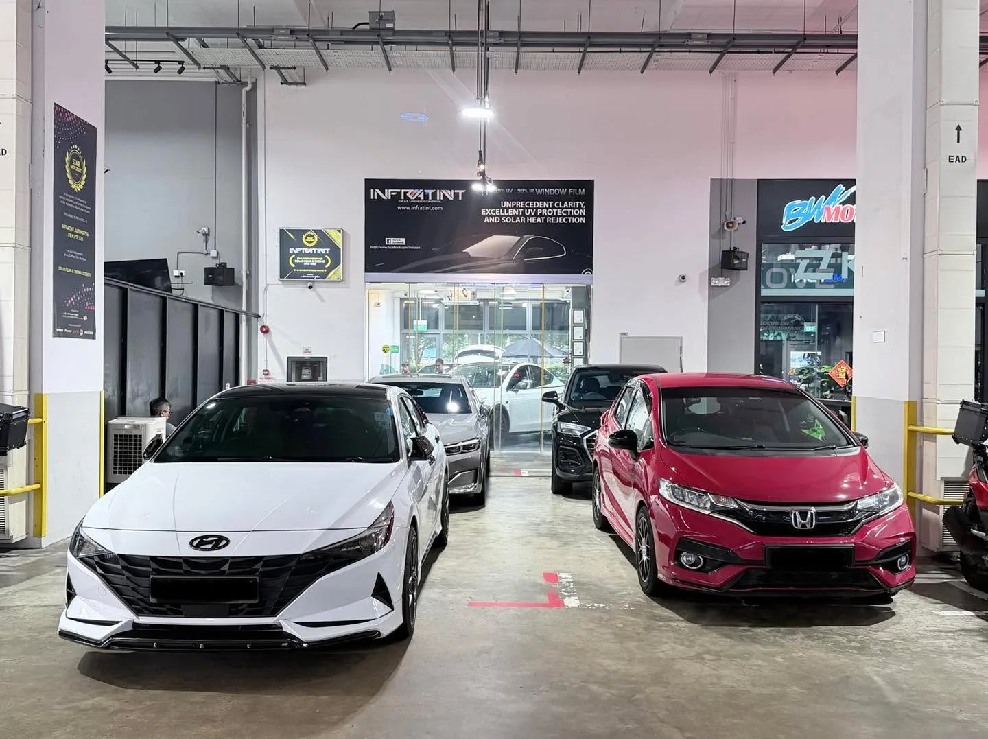 A white hatchback and a red hatchback parked indoors on grey flooring.
